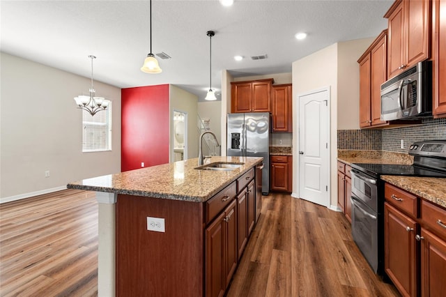 kitchen with decorative light fixtures, a sink, appliances with stainless steel finishes, brown cabinets, and a center island with sink