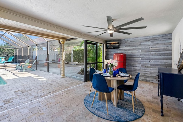 dining area with a sunroom, wooden walls, a ceiling fan, and a textured ceiling