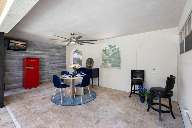 dining area with a textured ceiling, an accent wall, wood walls, a ceiling fan, and stone finish floor