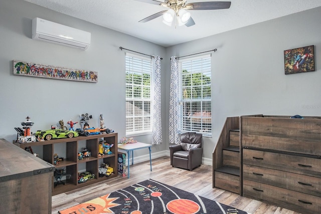 game room featuring a textured ceiling, a ceiling fan, baseboards, light wood-type flooring, and a wall mounted air conditioner