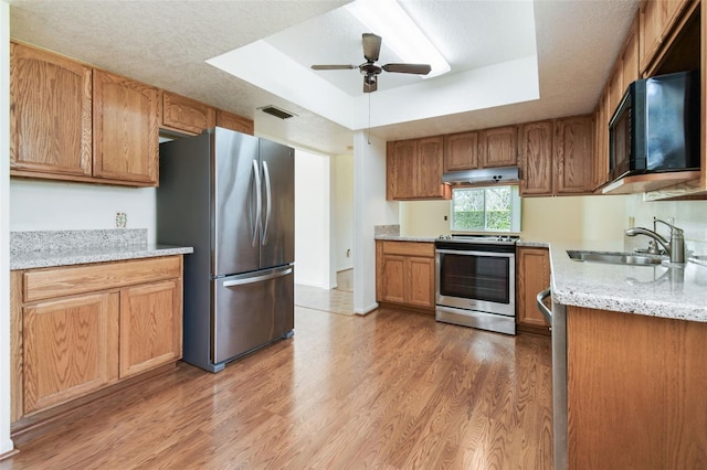 kitchen featuring a raised ceiling, light wood-style flooring, stainless steel appliances, under cabinet range hood, and a sink