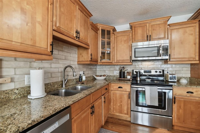 kitchen featuring brown cabinets, stainless steel appliances, glass insert cabinets, a sink, and light stone countertops