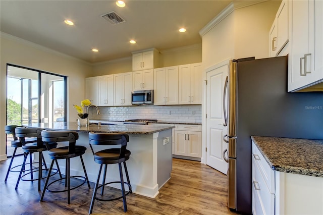 kitchen with stainless steel appliances, visible vents, white cabinetry, an island with sink, and dark stone countertops