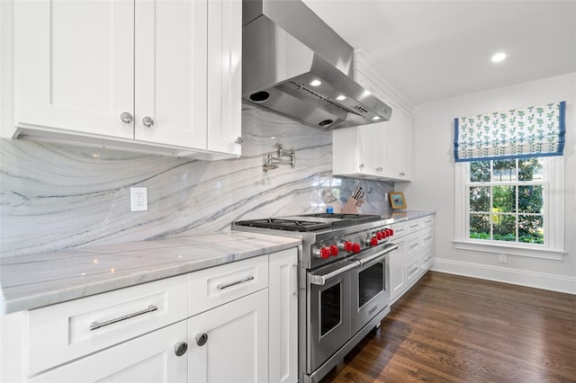 kitchen featuring range with two ovens, decorative backsplash, light stone countertops, wall chimney range hood, and white cabinetry