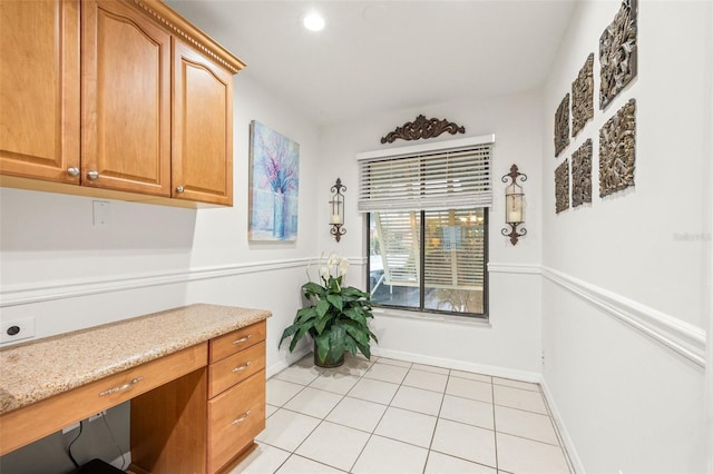 kitchen featuring light tile patterned floors, light stone counters, baseboards, brown cabinets, and built in desk