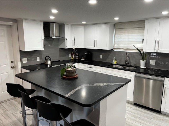 kitchen with white cabinets, dark countertops, wall chimney exhaust hood, stainless steel dishwasher, and a sink