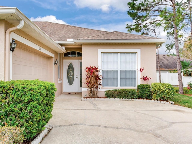 view of exterior entry featuring fence, concrete driveway, roof with shingles, stucco siding, and a garage