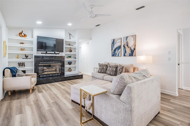 living room featuring built in features, a fireplace, light wood finished floors, ceiling fan, and baseboards