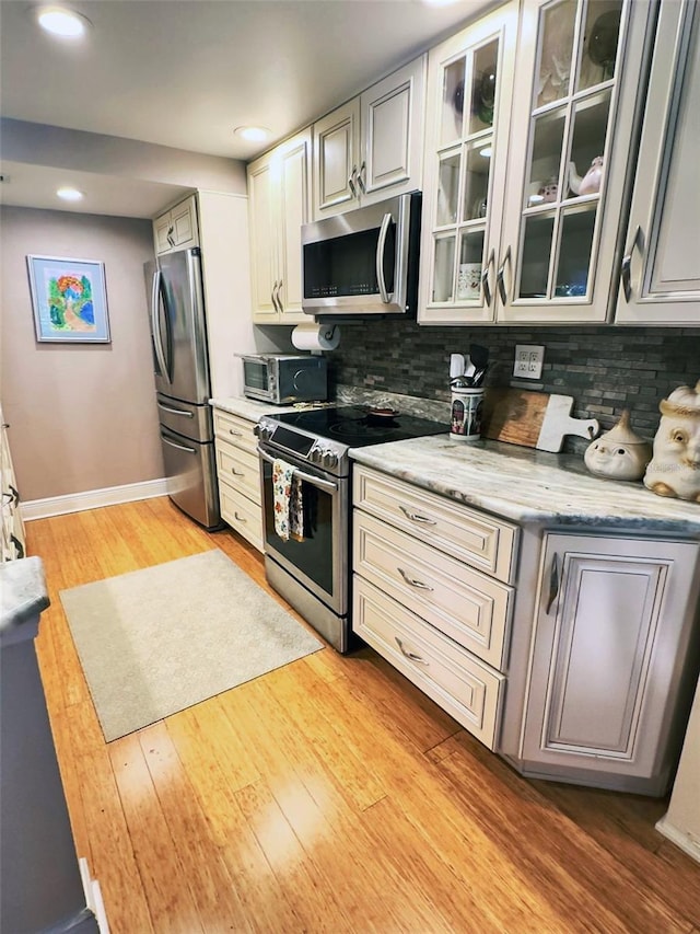 kitchen featuring tasteful backsplash, light wood-style flooring, appliances with stainless steel finishes, glass insert cabinets, and baseboards