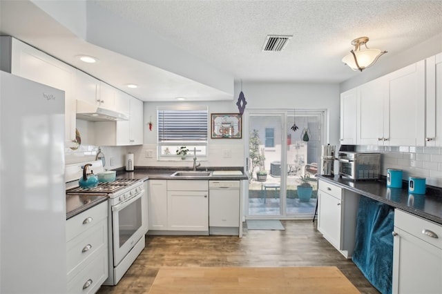 kitchen featuring visible vents, a sink, under cabinet range hood, dark countertops, and white appliances