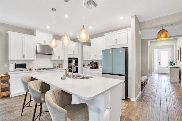 kitchen featuring under cabinet range hood, a sink, appliances with stainless steel finishes, backsplash, and an island with sink