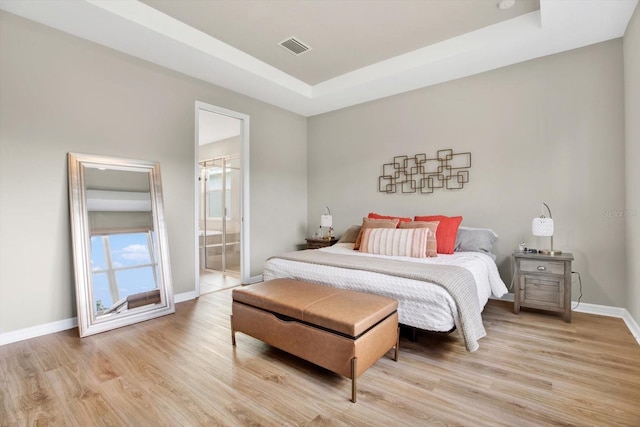 bedroom with light wood finished floors, baseboards, visible vents, a raised ceiling, and connected bathroom