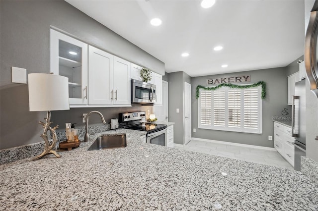 kitchen with light stone counters, a sink, white cabinetry, appliances with stainless steel finishes, and glass insert cabinets