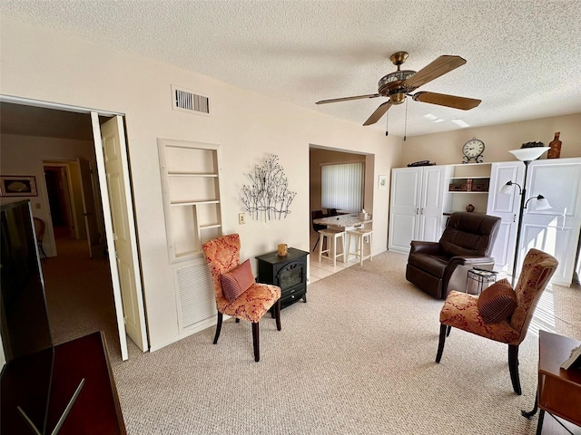 living area featuring ceiling fan, a textured ceiling, light carpet, visible vents, and a wood stove