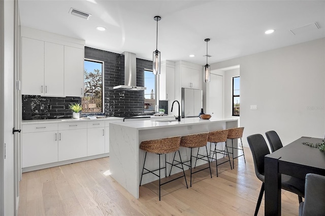 kitchen featuring visible vents, fridge, a kitchen island with sink, wall chimney range hood, and pendant lighting