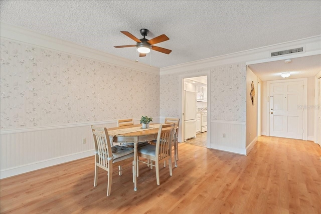 dining space with a textured ceiling, wainscoting, wallpapered walls, light wood finished floors, and crown molding