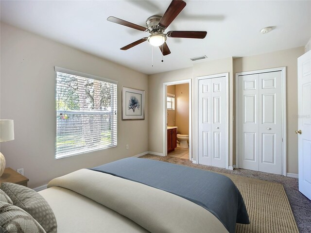 bedroom featuring two closets, light colored carpet, visible vents, ensuite bathroom, and baseboards