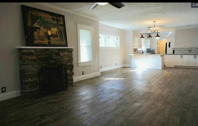 unfurnished living room featuring baseboards, dark wood finished floors, ornamental molding, a fireplace, and ceiling fan with notable chandelier