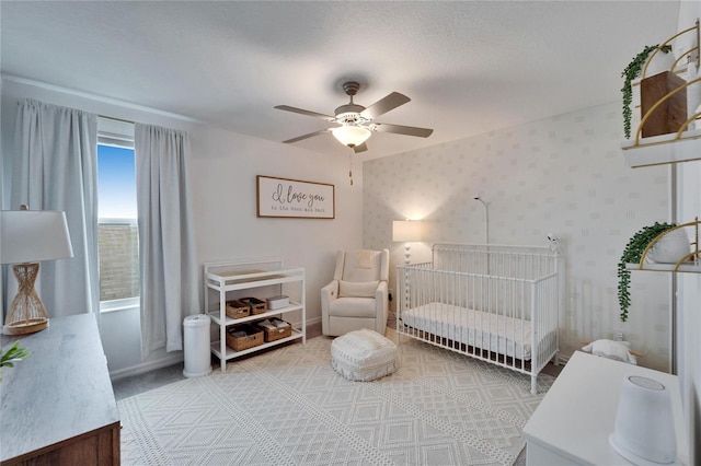 bedroom featuring a crib, wallpapered walls, baseboards, a ceiling fan, and a textured ceiling