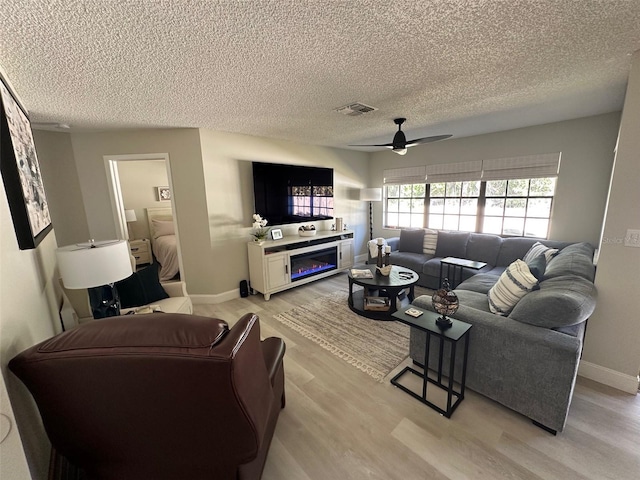 living area with visible vents, a glass covered fireplace, ceiling fan, a textured ceiling, and light wood-style floors