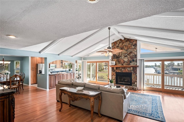 living area featuring light wood-style flooring, lofted ceiling with beams, a stone fireplace, a textured ceiling, and ceiling fan with notable chandelier