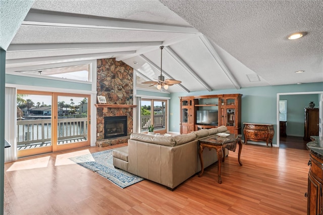 living room featuring vaulted ceiling with beams, light wood finished floors, ceiling fan, a stone fireplace, and a textured ceiling