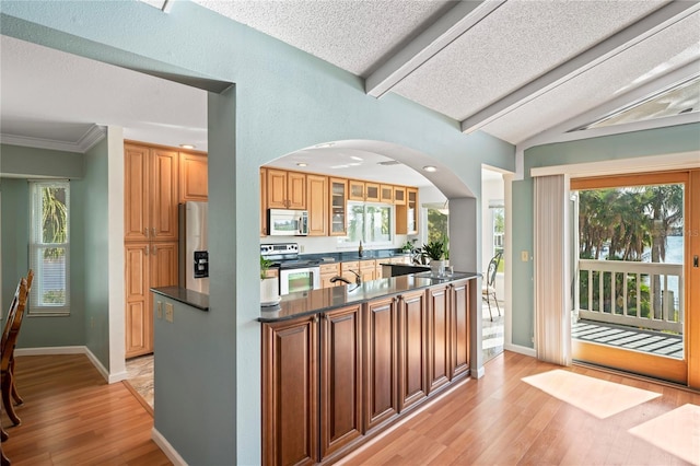 kitchen featuring arched walkways, vaulted ceiling with beams, light wood-style flooring, stainless steel appliances, and dark countertops