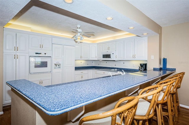 kitchen featuring a breakfast bar, dark wood finished floors, white appliances, a peninsula, and a raised ceiling