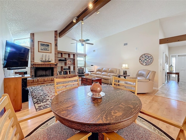 dining area with a textured ceiling, light wood-type flooring, a brick fireplace, a wealth of natural light, and beamed ceiling