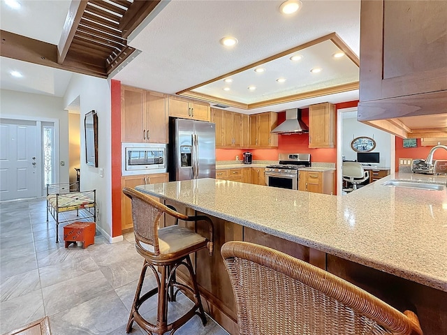 kitchen featuring a peninsula, a tray ceiling, stainless steel appliances, wall chimney range hood, and a sink