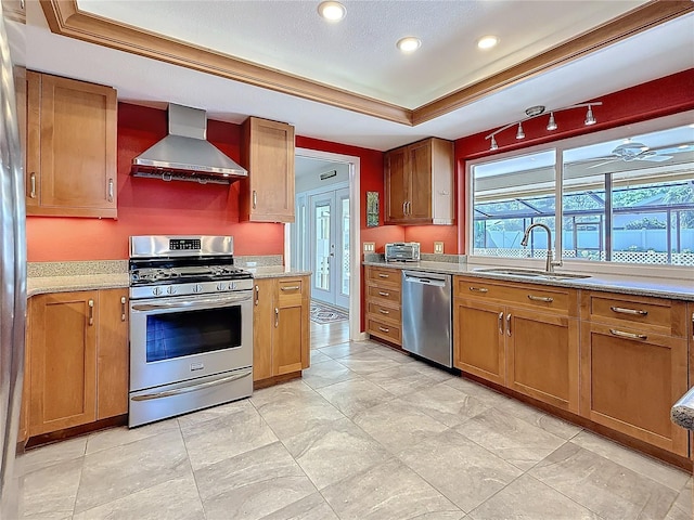 kitchen featuring a raised ceiling, appliances with stainless steel finishes, brown cabinets, wall chimney range hood, and a sink