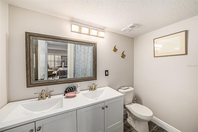 ensuite bathroom with visible vents, toilet, a sink, a textured ceiling, and double vanity