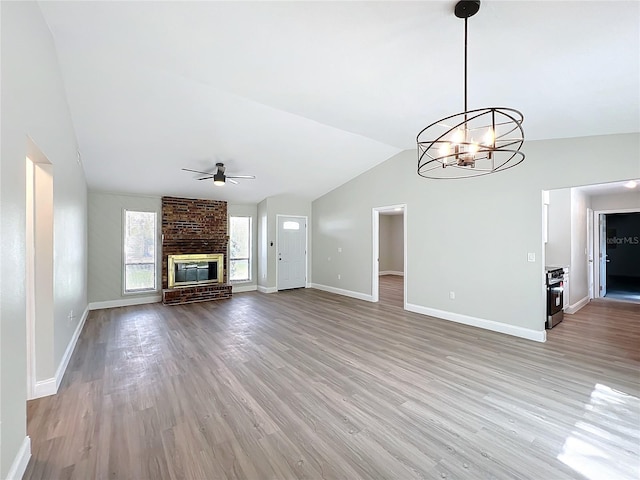 unfurnished living room with a brick fireplace, baseboards, lofted ceiling, light wood-style floors, and ceiling fan with notable chandelier