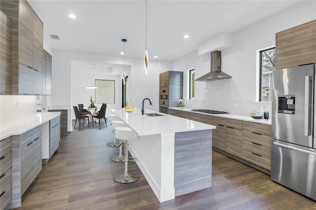 kitchen featuring stainless steel appliances, light countertops, wall chimney exhaust hood, modern cabinets, and a center island with sink