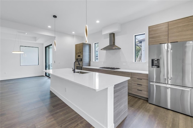 kitchen featuring stainless steel appliances, light countertops, wall chimney range hood, brown cabinetry, and a center island with sink