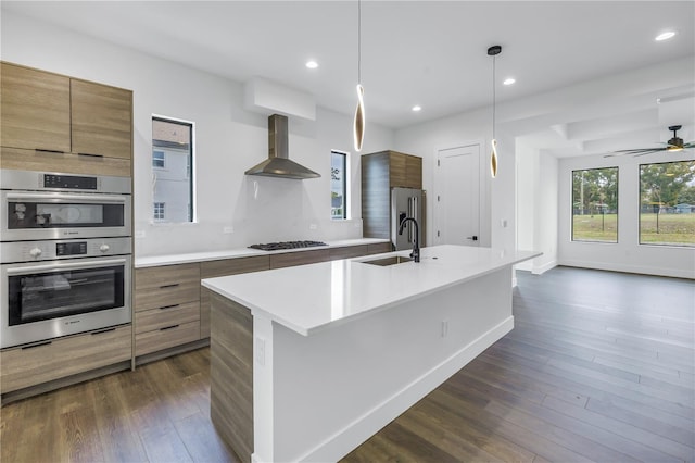 kitchen featuring stainless steel appliances, light countertops, brown cabinetry, a kitchen island with sink, and wall chimney range hood