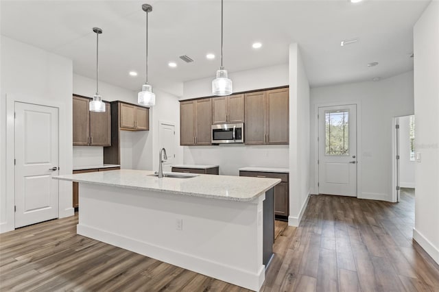 kitchen featuring light stone counters, pendant lighting, stainless steel microwave, a sink, and an island with sink