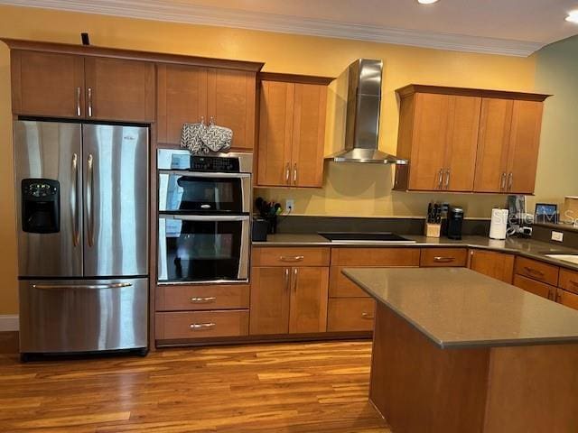 kitchen featuring brown cabinetry, appliances with stainless steel finishes, ornamental molding, wood finished floors, and wall chimney range hood