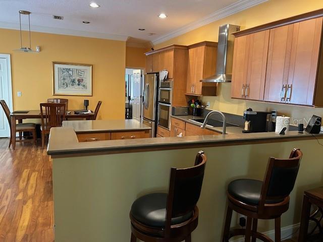 kitchen with crown molding, light wood finished floors, stainless steel appliances, brown cabinetry, and wall chimney range hood