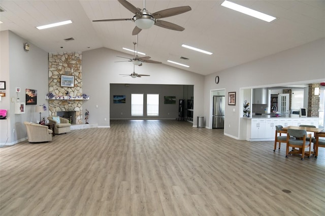 living room with light wood-style floors, ceiling fan, a stone fireplace, high vaulted ceiling, and baseboards