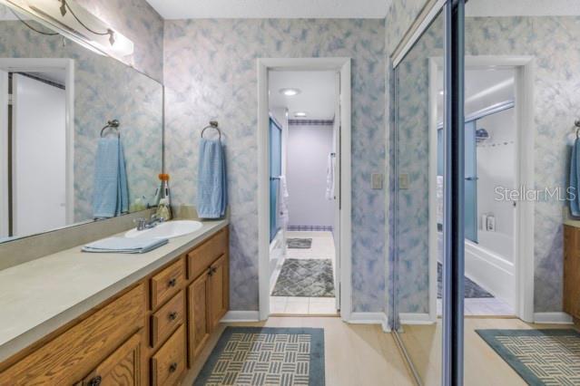 bathroom featuring tile patterned flooring, shower / bath combination with glass door, vanity, baseboards, and wallpapered walls