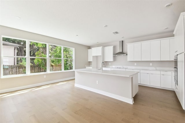 kitchen featuring an island with sink, light countertops, light wood-type flooring, wall chimney range hood, and a sink