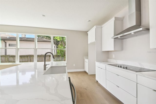 kitchen featuring wall chimney exhaust hood, light stone counters, black electric cooktop, white cabinetry, and a sink