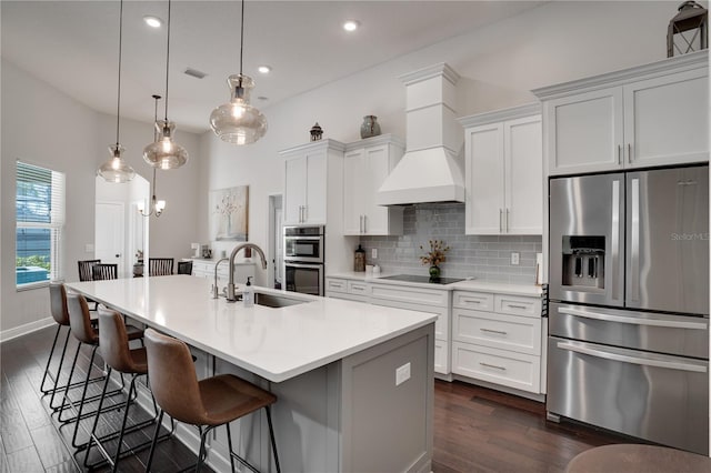 kitchen featuring an island with sink, backsplash, custom exhaust hood, stainless steel appliances, and a sink