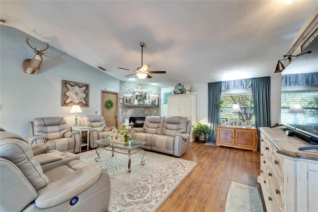 living room with a stone fireplace, light wood-style flooring, visible vents, a ceiling fan, and vaulted ceiling