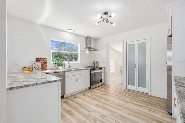 kitchen with white cabinets, wall chimney exhaust hood, appliances with stainless steel finishes, light stone countertops, and light wood-style floors