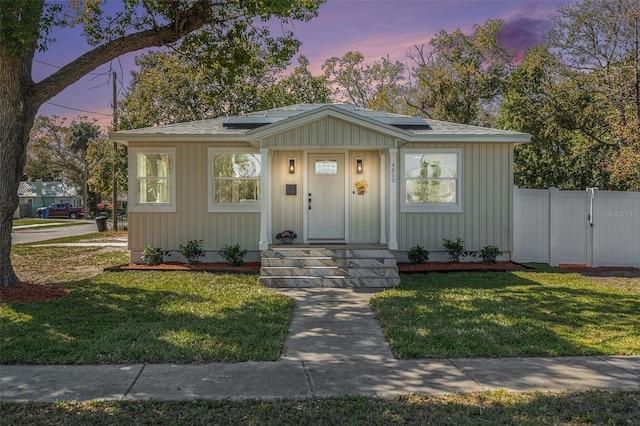 bungalow-style home featuring solar panels, a shingled roof, board and batten siding, fence, and a front lawn