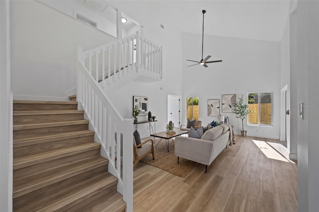 living area featuring light wood-style flooring, a high ceiling, visible vents, a ceiling fan, and stairs