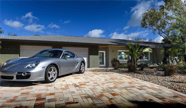 ranch-style home featuring an attached garage, a shingled roof, decorative driveway, and stucco siding