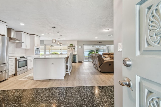 kitchen featuring appliances with stainless steel finishes, open floor plan, white cabinets, a textured ceiling, and wall chimney exhaust hood